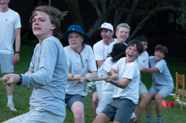 A group of boys in casual attire engage in a tug-of-war game outdoors, displaying varying expressions of effort and enjoyment.