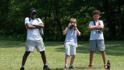 Three children in casual clothes stand in a grassy field. One is taking a photo with a camera, and the other two have their arms crossed.
