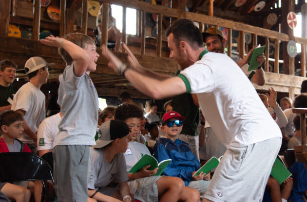 A man and a boy exchange a high-five amidst a group of seated children inside a rustic building, all appearing to be engaged in a lively group activity or event.