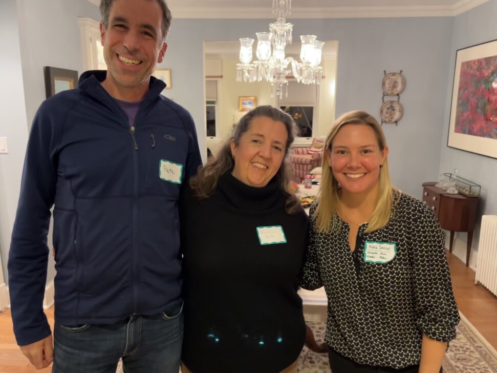 Three people stand indoors, each wearing a name tag, smiling at the camera. The background includes a chandelier, a table with food, and wall decorations.
