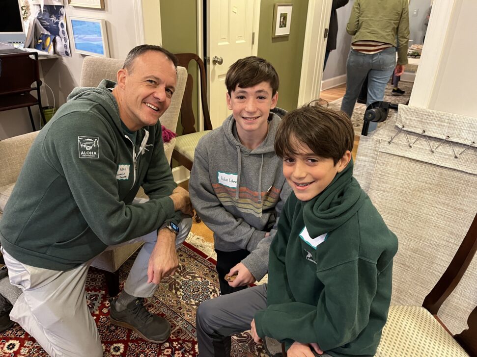 A man and two young boys, all wearing name tags, are sitting and smiling in a room with a rug and beige walls.