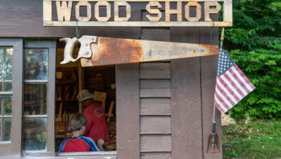 Exterior view of a wood shop with a sign, a rusty saw hanging, and an American flag.