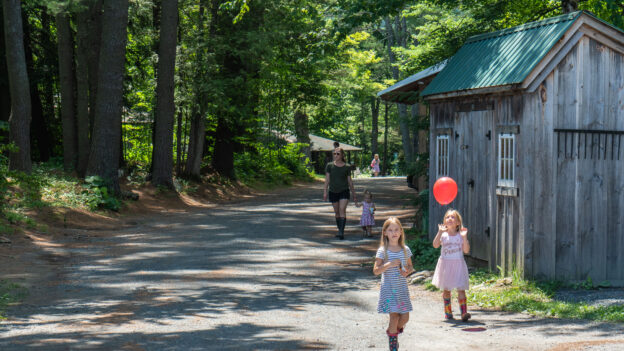 Children walk along a forested path near a rustic wooden building, with one child holding a red balloon, while an adult follows behind.