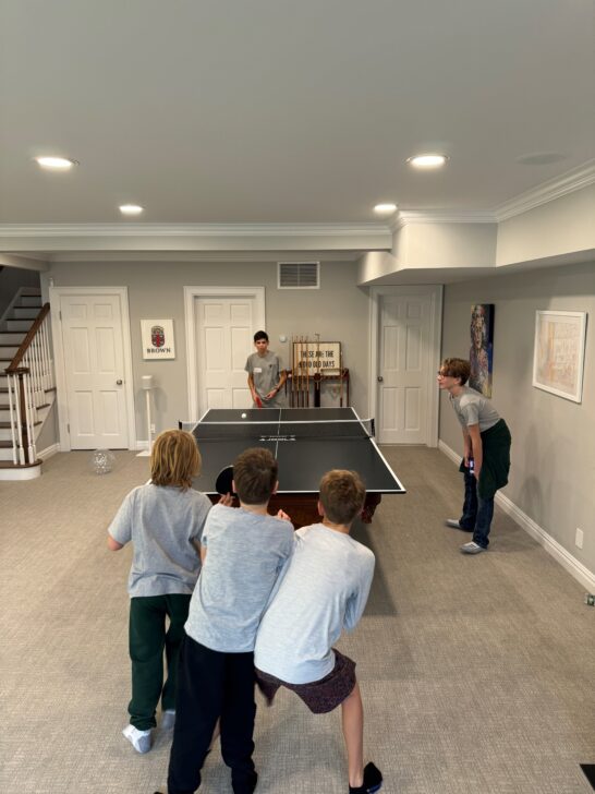 Five boys are in a room playing a game of table tennis. Four boys, on the left side, are watching one boy serve the ball.