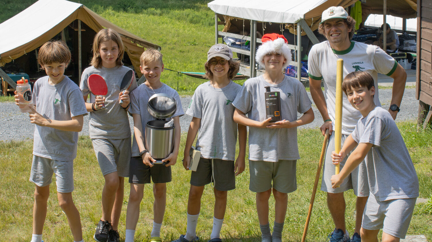 A group of five boys and one man stand outdoors, smiling. The boys hold various items, including a ping pong paddle, trophy, box, and a broom.