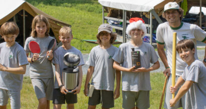 A group of five boys and one man stand outdoors, smiling. The boys hold various items, including a ping pong paddle, trophy, box, and a broom.