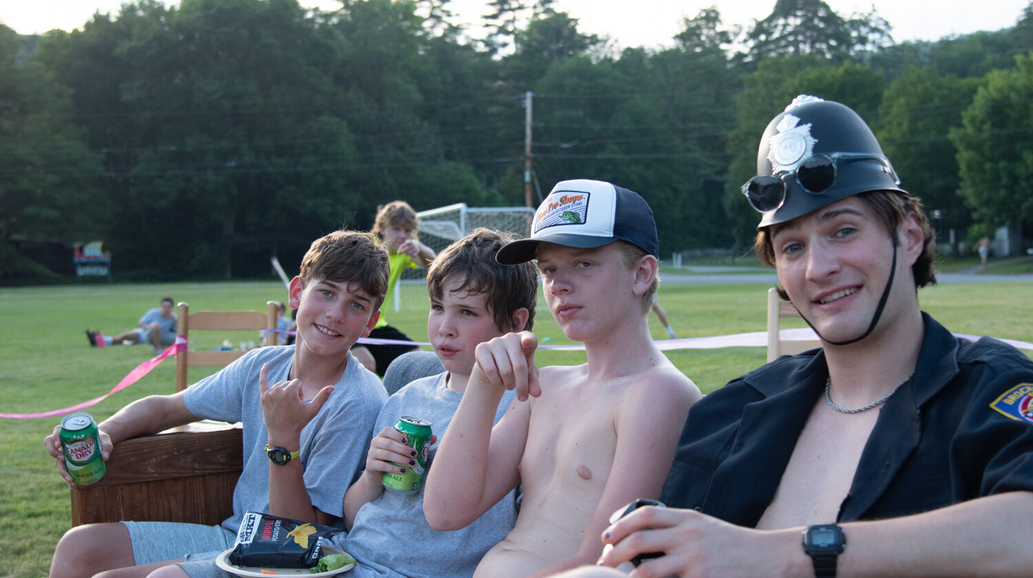 Four boys seated outdoors on a bench. The boy on the left holds a drink and makes a hand gesture, while the others hold beverage cans.