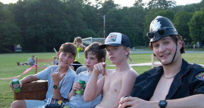Four boys seated outdoors on a bench. The boy on the left holds a drink and makes a hand gesture, while the others hold beverage cans.